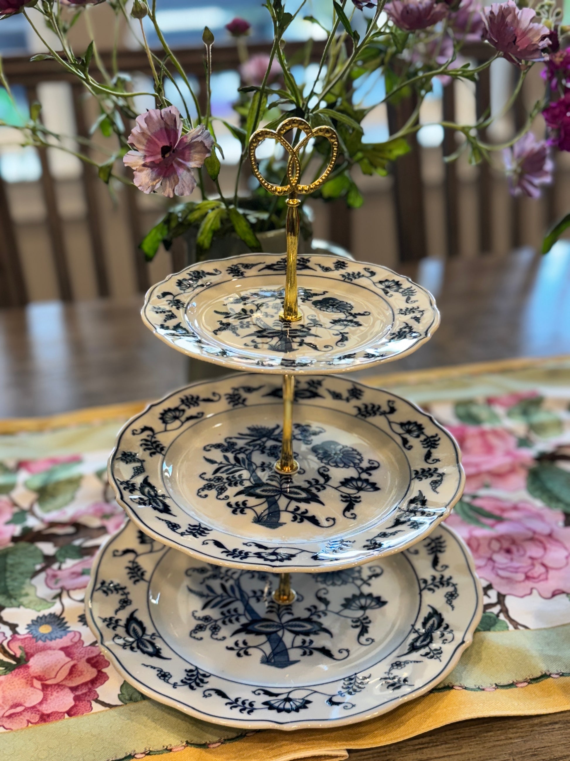 Three-tiered stand with blue onion floral porcelain plates and gold heart handle, displayed on floral runner with soft lighting
