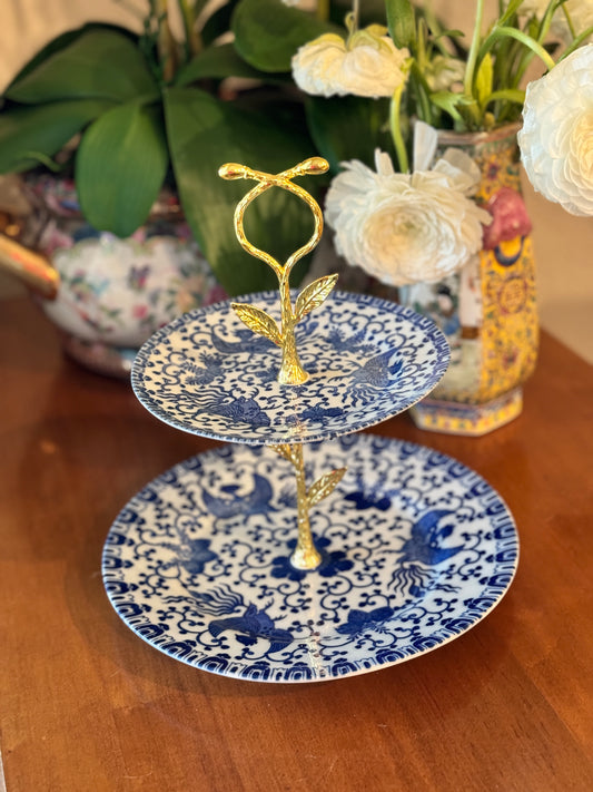 Two-tiered stand with blue and white porcelain plates featuring bird and scroll design, gold floral handle, styled on wood table with floral background