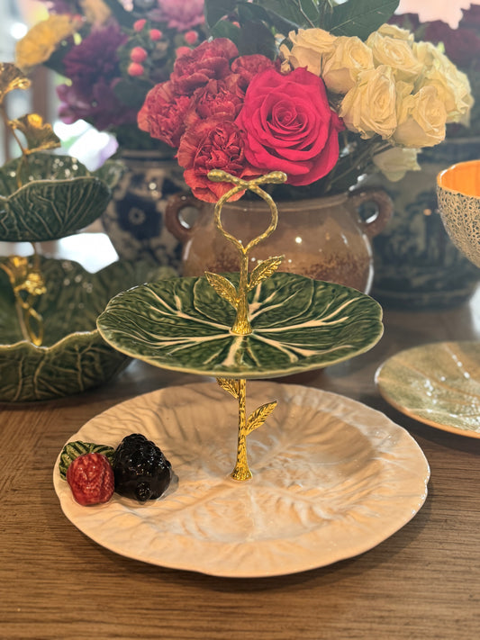 Two-tiered stand made from green and cream cabbage leaf plates with gold leaf hardware, styled with fruit and floral centerpiece on wood table