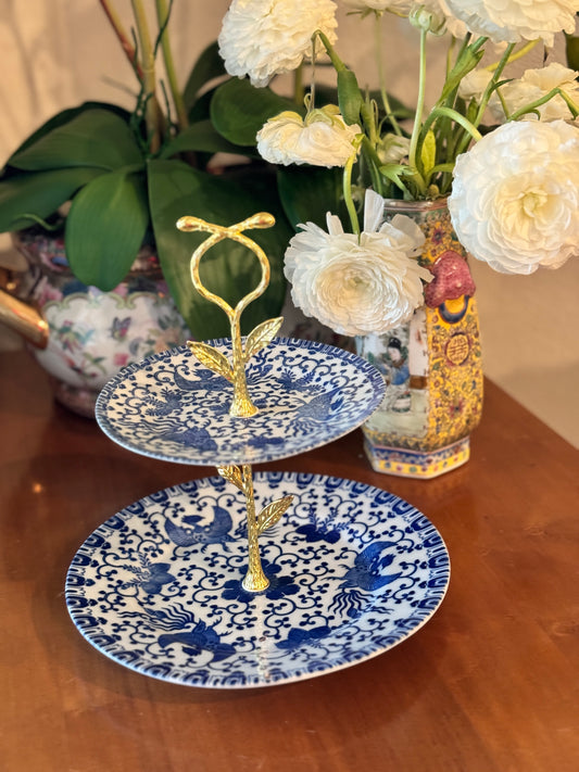 Two-tiered stand with blue and white porcelain plates featuring bird and scroll design, gold floral handle, styled on wood table with floral background
