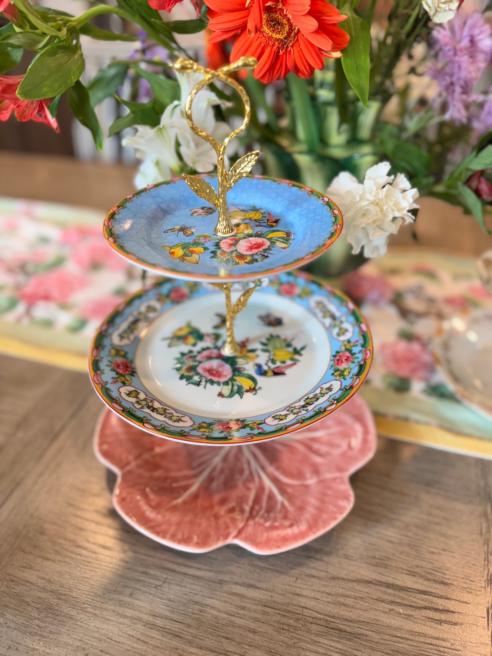 Three-tiered stand made from vintage floral china with gold hardware, featuring a pink cabbage leaf base, Famille Rose middle plate, and blue floral top plate, surrounded by fresh flowers on a wooden table.