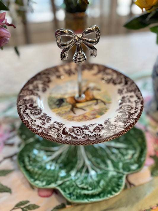 Two-tiered stand with a Spode Woodland American Quarter horse plate on top and a green Bordallo cabbage leaf plate on the bottom, featuring a silver bow handle, styled on a floral table runner.