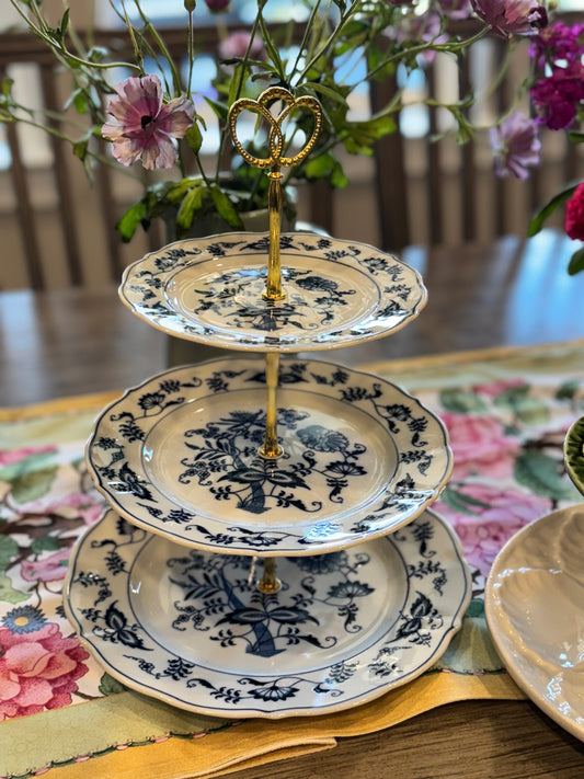 Three-tiered stand with blue onion floral porcelain plates and gold heart handle, displayed on floral runner with soft lighting
